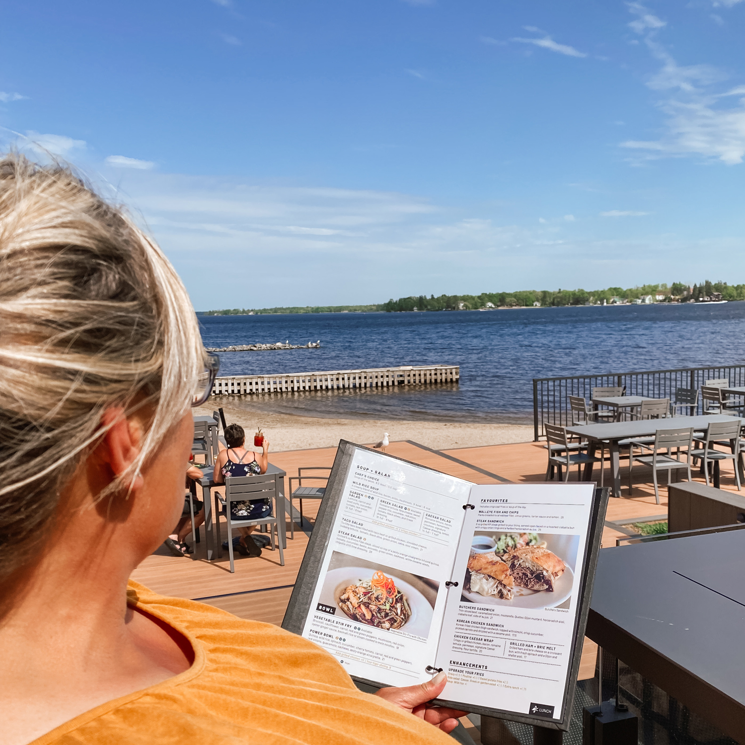 a woman relaxes and holds up a menu on a large, breezy outdoor patio overlooking a beach, dock and a wide blue lake on a summer day. 