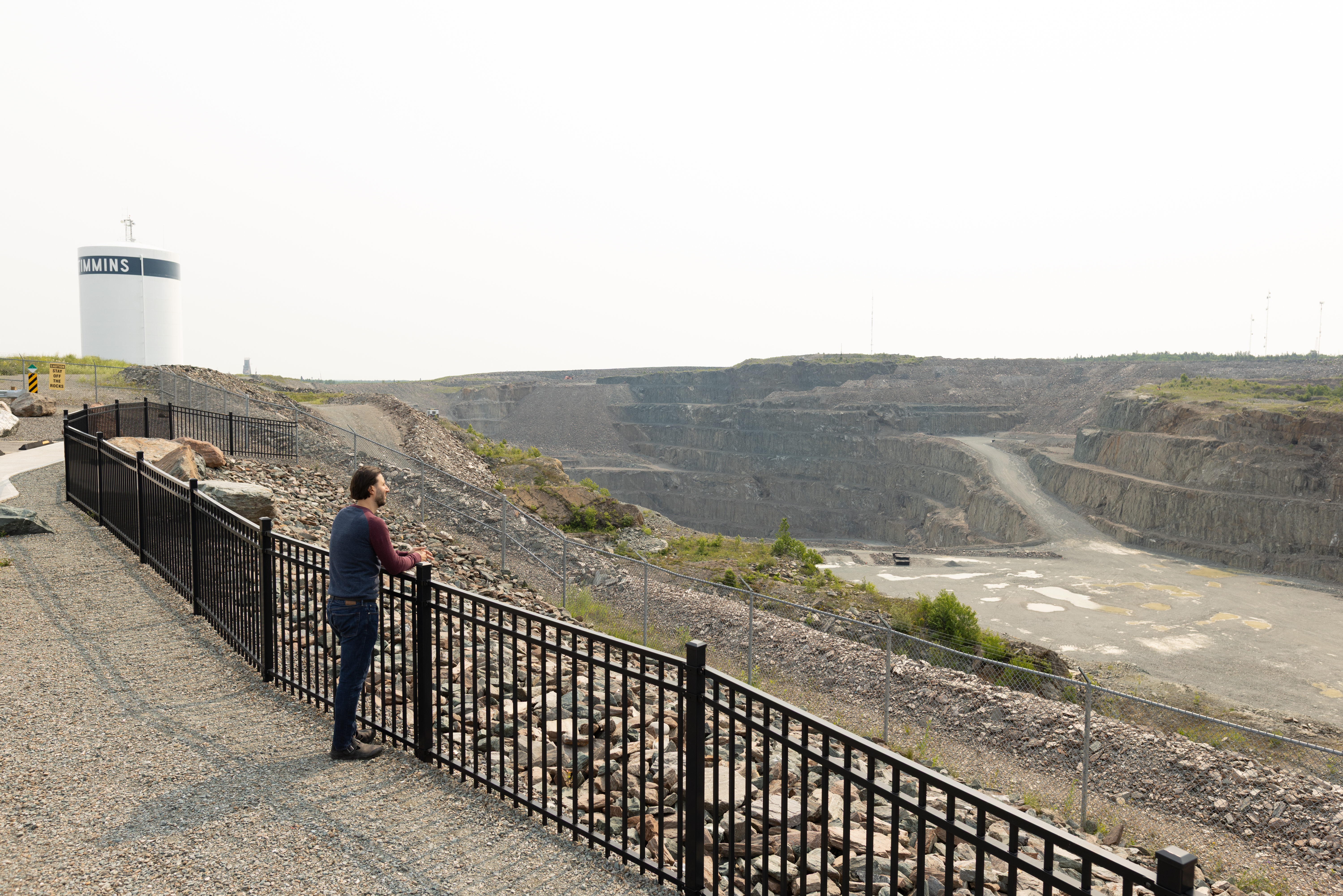 a person looks out over the Hollinger Open Pit Mine, standing with their hands rested on the black iron fence that surrounds it. The Timmins water tower is in the background. 