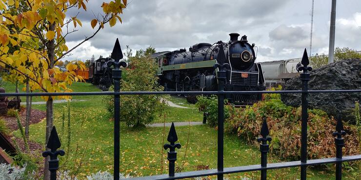 a vintage iron locomotive sits in a treed park area surrounded by a wrought iron fence in the autumn at the Northern Ontario Railroad Museum in Capreol. 
