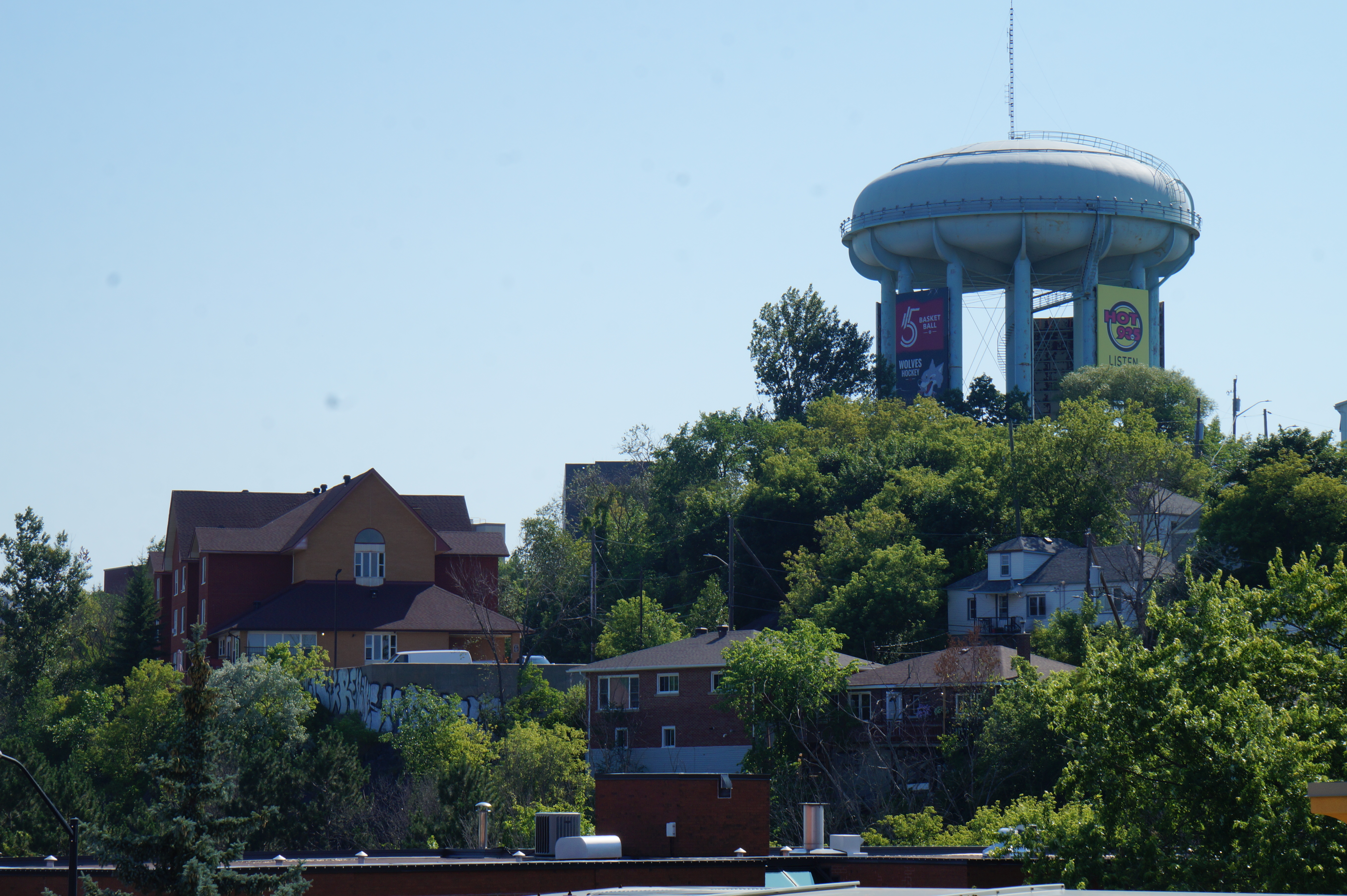 The Sudbury watertower stands shining on a sunny summer day, towering above stately homes tucked among clusters of lush green trees.