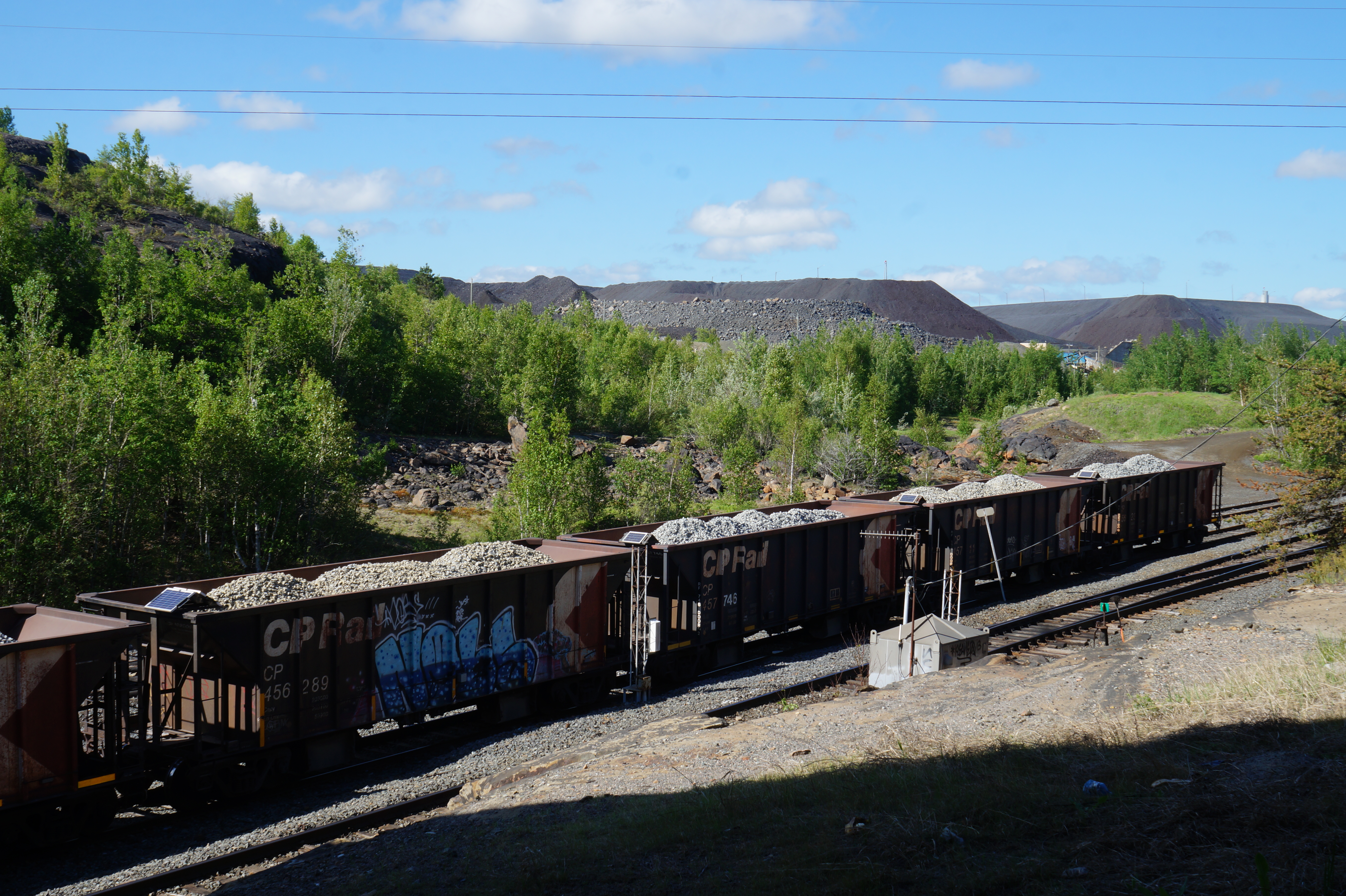 loaded coal cars on a train track near Sudbury, surrounded by green forest, low mountains and bright blue sky. 