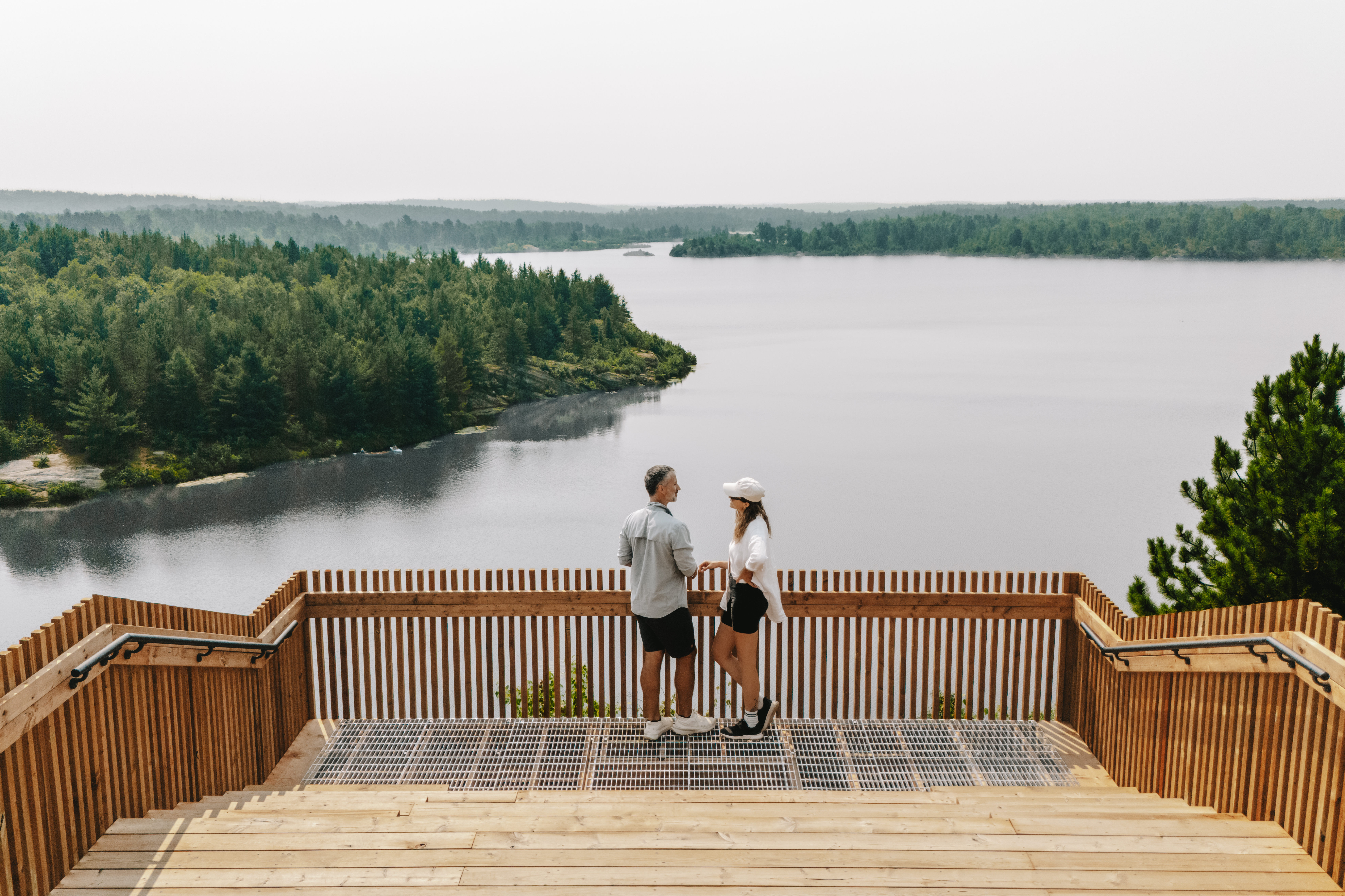 Two smiling people talk as they stand on a wooden platform looking out over a shining lake and green forest at Lake Laurentian Conservation Area. 