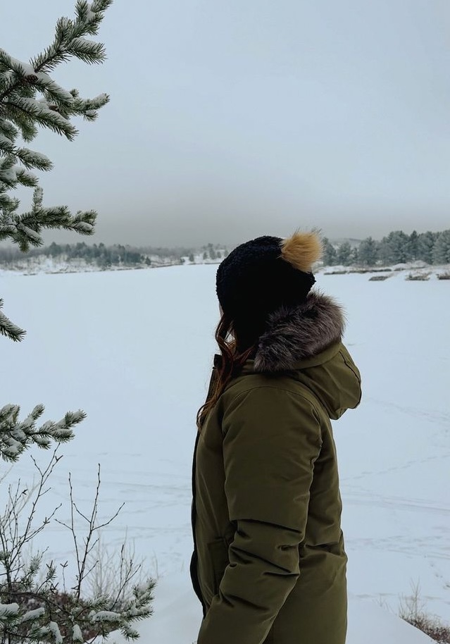 a woman bundled up against the cold looks out over a frozen lake from a forest clearing on a grey winter day at Lake Laurentian Conservation Area. 