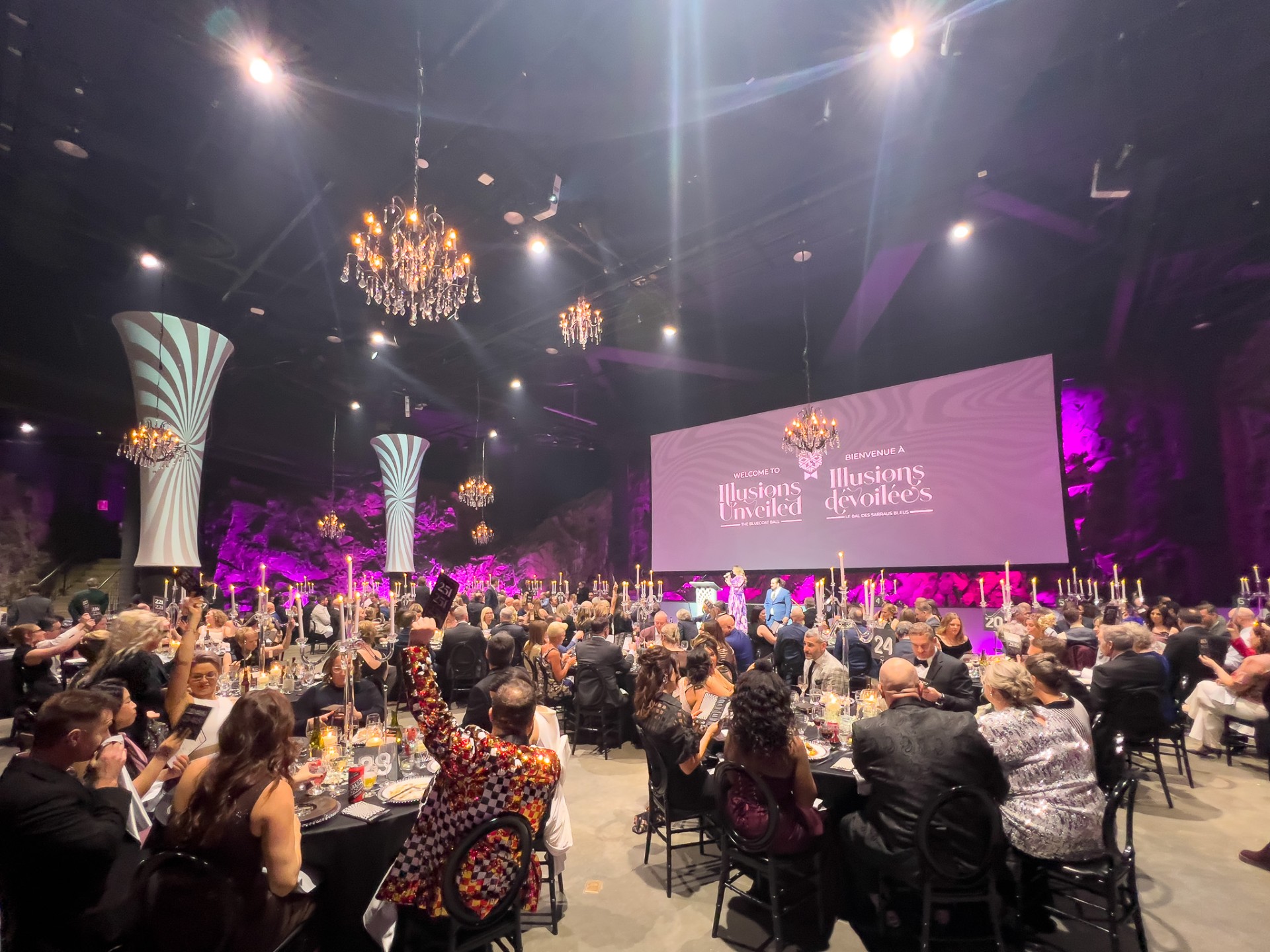 tables of conference-goers, many with raised hands, look at an illuminated screen at the end of the huge, lavishly decorated rock cavern meeting room at Science North in Sudbury. 