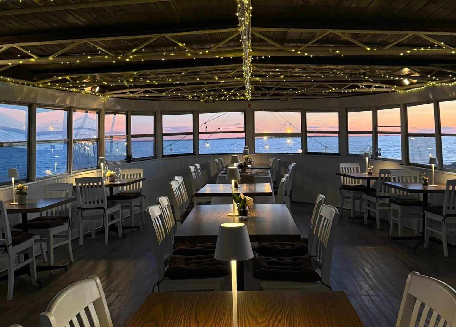 rows of white chairs and wooden tables set with softly glowing lamps under white twinkle lights inside a wooden reception room at The Boat in North Bay. Windows in all directions show a soft pink and blue sunset glowing outside.