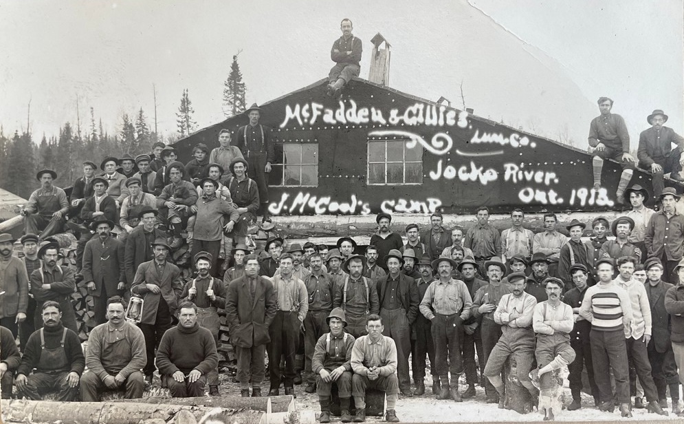 a historical photograph of a camp full of logging company workers of the McFadden & Gillies Lumber Company posing in front of their camp building at Jocko River in 1913.