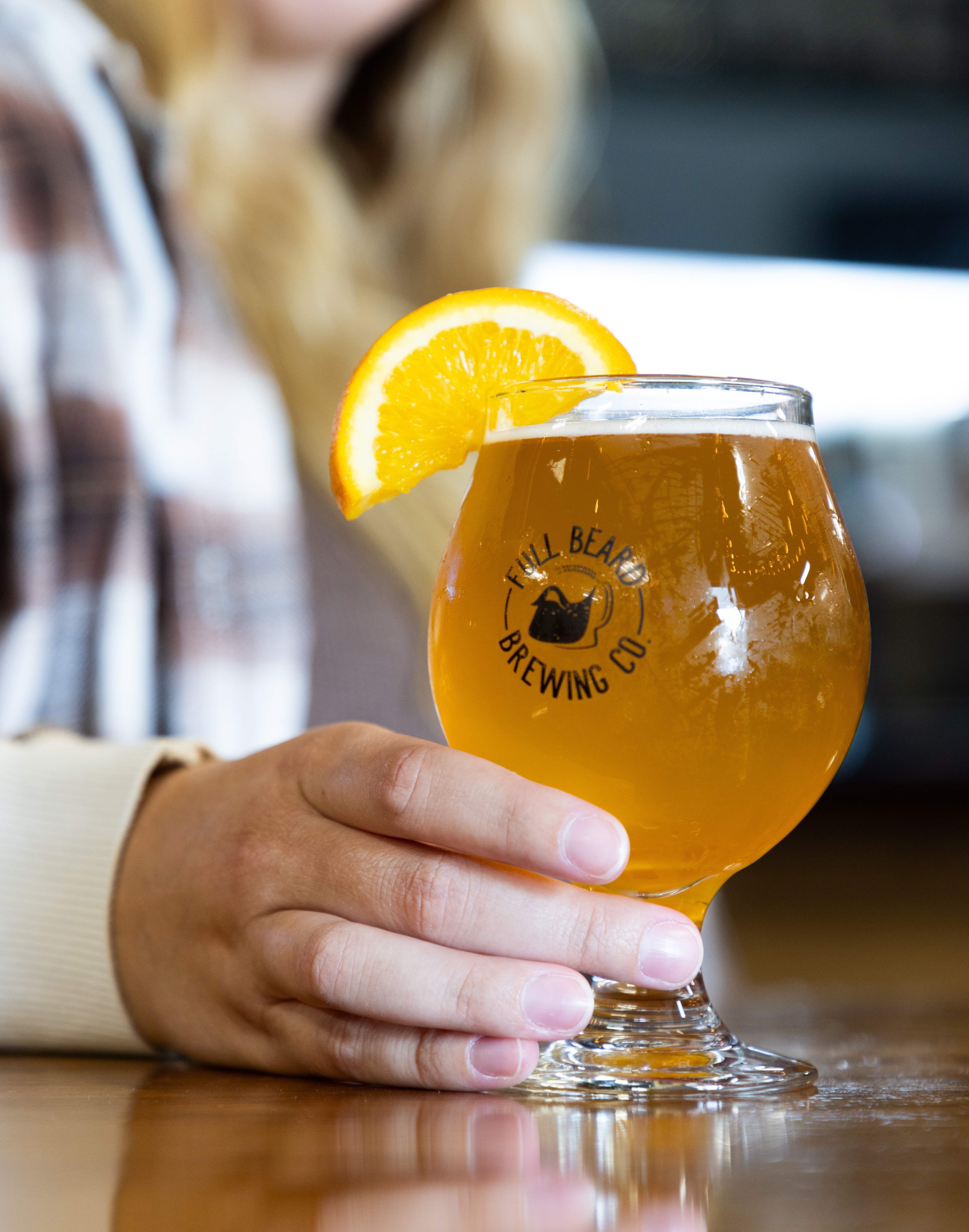 a person holding a glass of Full Beard Brewing Company beer adorned with a lemon wedge. 