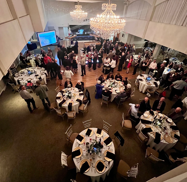 At the Porcupine Dante Club, circular tables decorated elegantly for dinner fill a large hall filled with mingling people under large crystal chandeliers.
