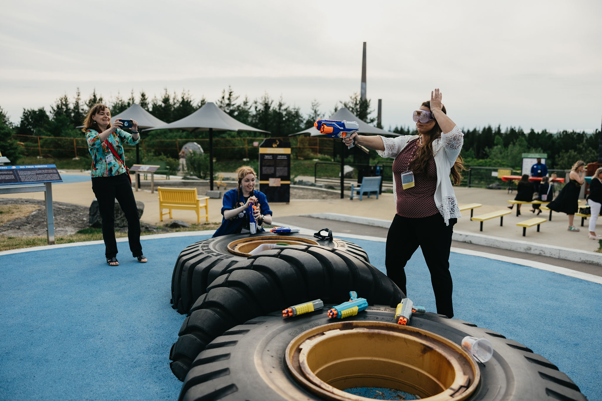 A delegate wearing a conference lanyard around her neck poses as she shoots a NERF pistol at an obstacle course at Dynamic Earth as a co-worker take a video and smiling Science North staff looks on.