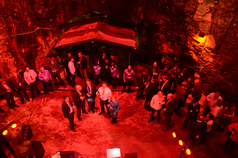 conference attendees sip drinks and mingle along the rock walls of the Vale Cavern under red lights. 