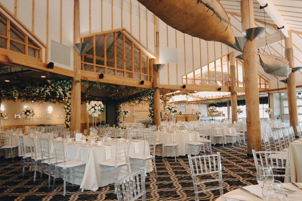 White wooden chairs and tables laid elegantly for dinner in a dining room at McGillivray's Landing, with large, high windows and ceilings. Birchbark canoes are suspended from the rafters.