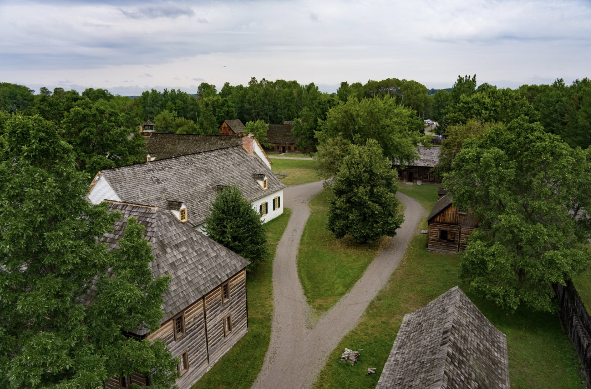 Wooden historical buildings along a forked path tucked between large, lush green trees at Fort William Historical Park.