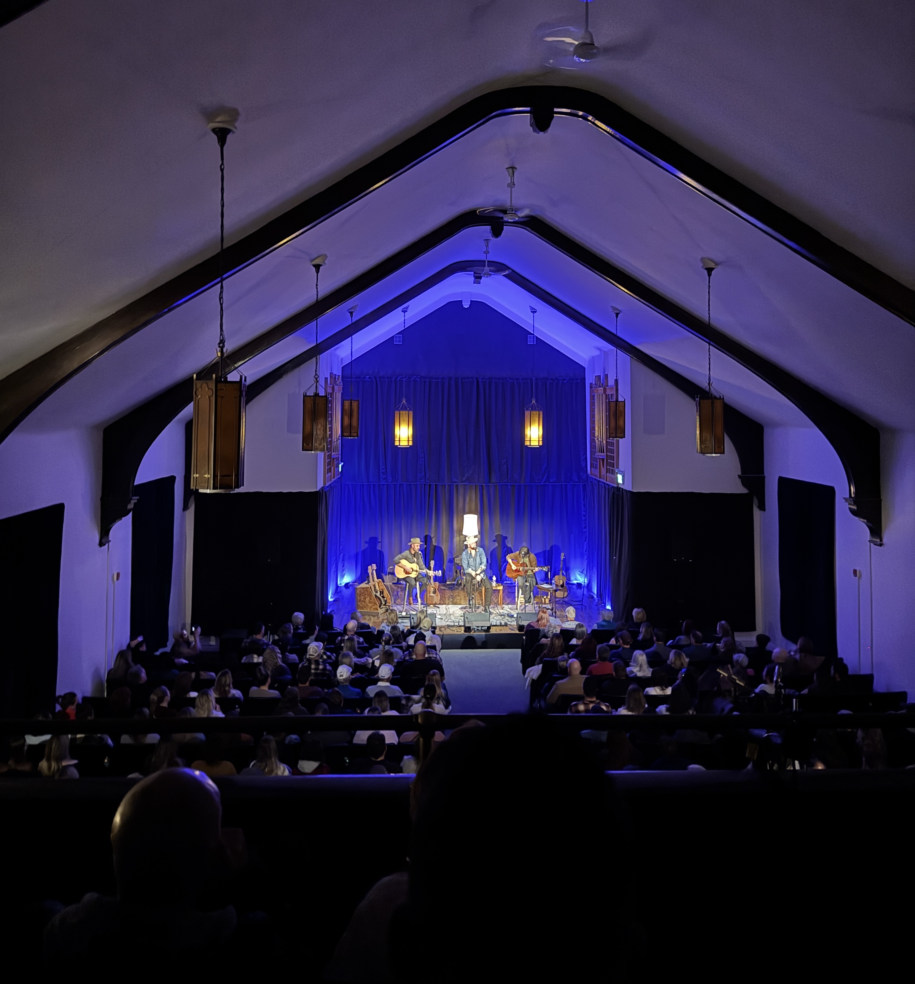 a band plays on the small blue-lit stage at Knox Hall in front of a full audience, all dwarfed under the high peaked ceiling.
