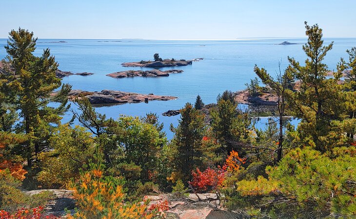 the glassy blue water of Lake Huron stretches to the horizon, dotted with large smooth rocky shoals and surrounded by bright red, green and gold autumn trees. 