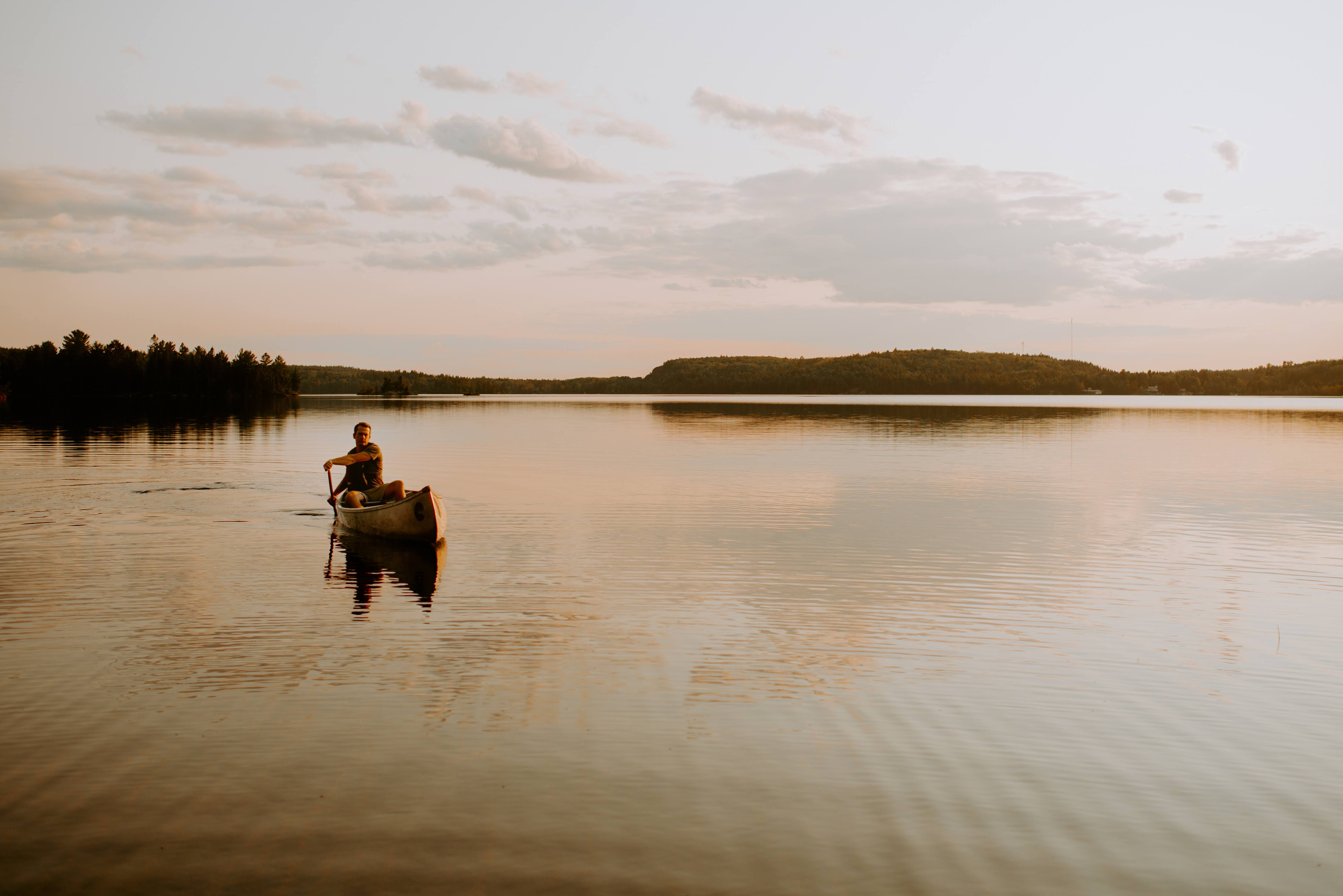 a person paddles a canoe on the glassy calm lake in Sudbury, the water reflecting the soft golden sunset above.