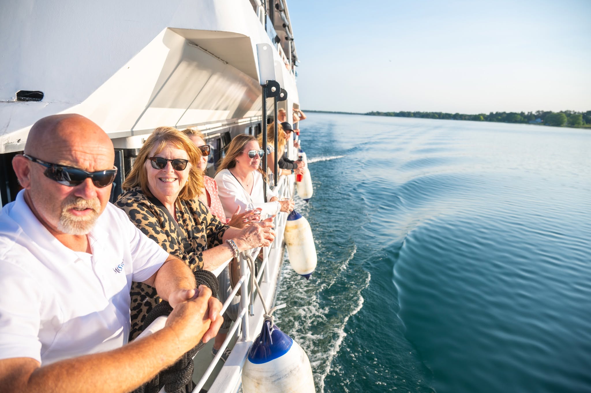 smiling passengers in sunglasses lined up along the rail of the Miss Marie tour boat, looking out over the blue shining water as they sail along on a crystal clear sunny day in Sault Ste. Marie. 