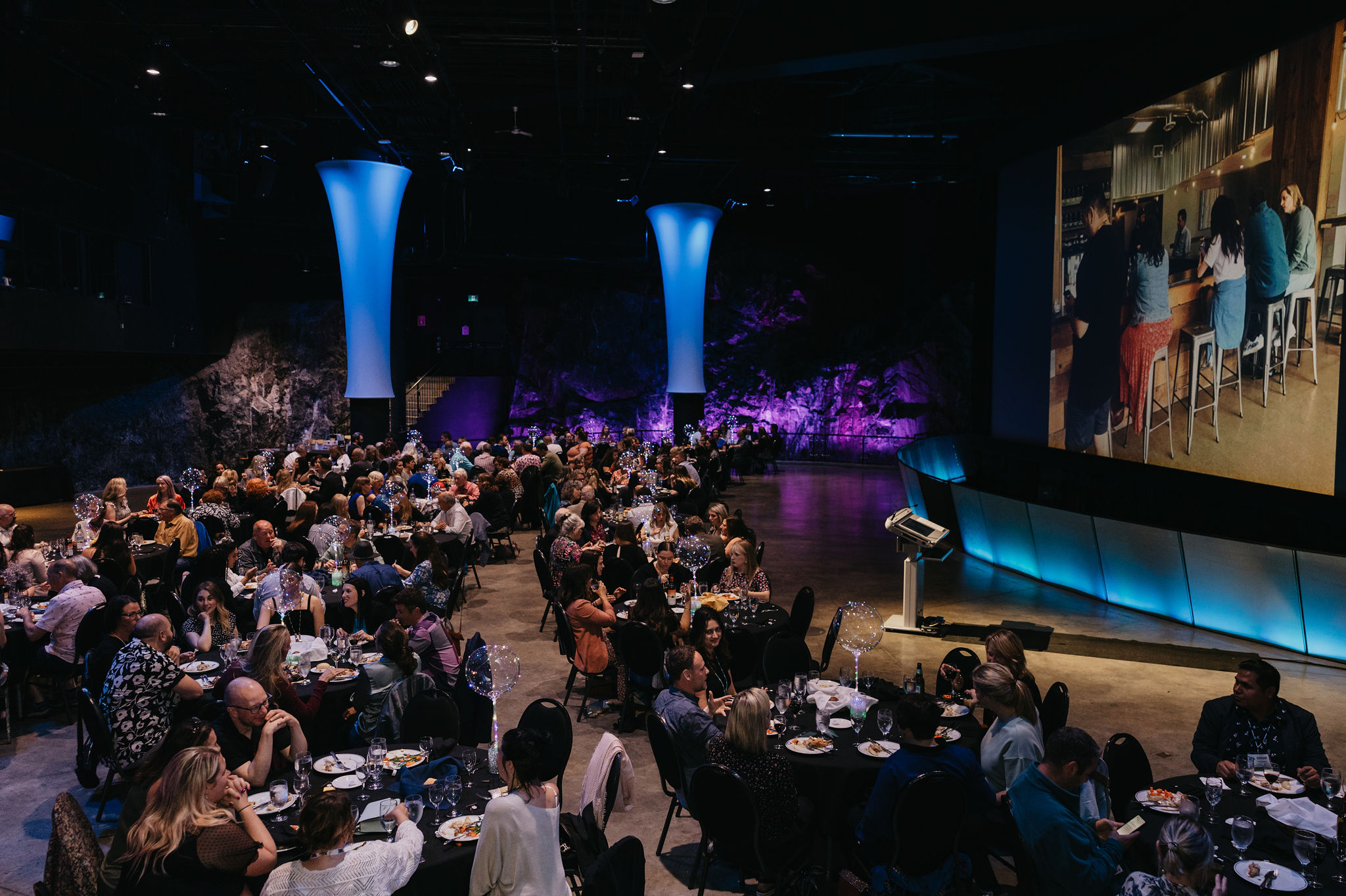 The Vale Cavern at Science North, filled with conference attendees watching a tall presentation screen. Blue lighted pillars softly illuminate the rough rock walls of the cavern in the background.
