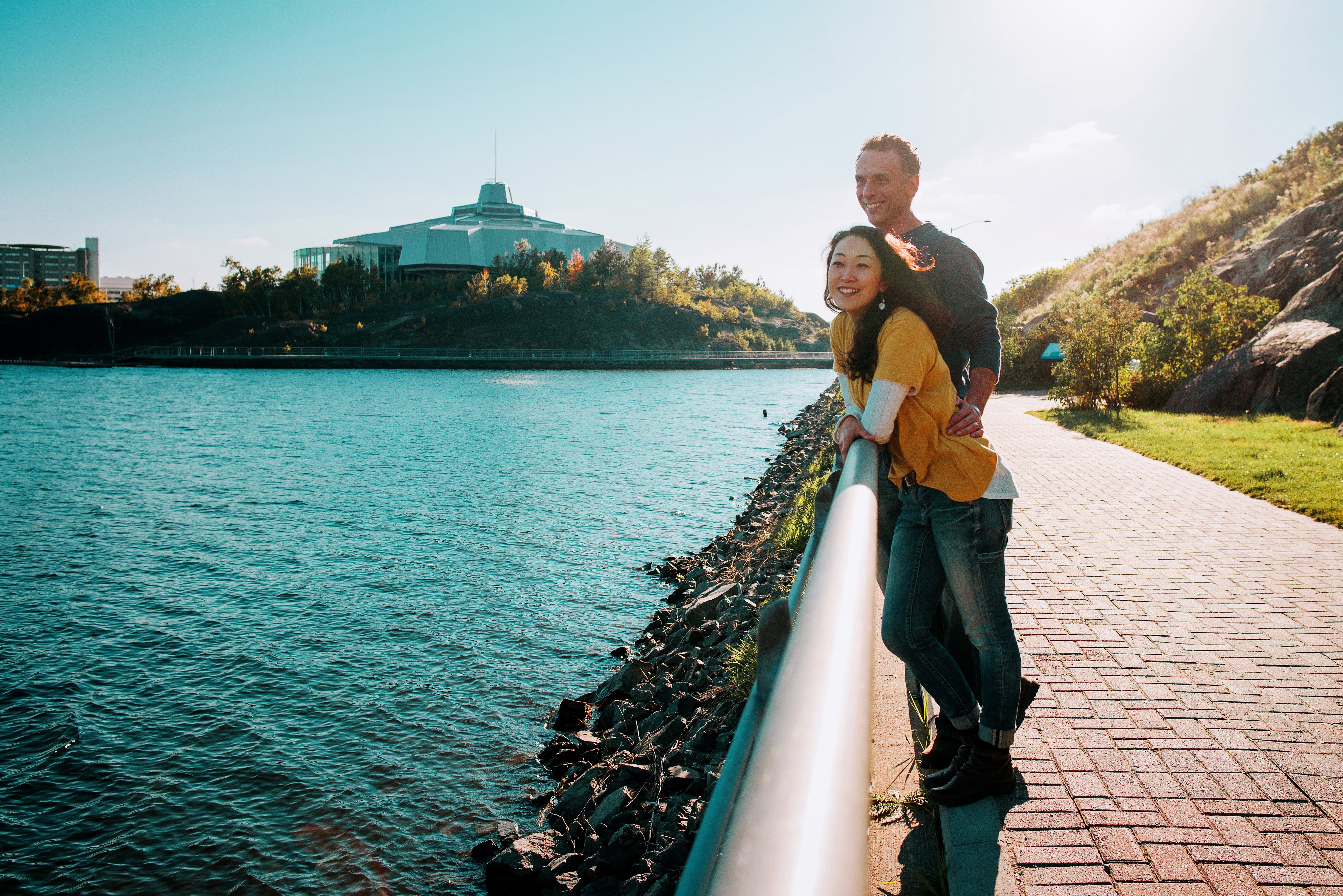 two people look out over a rail along the boardwalk at the shining blue Lake Ramsey on a clear, breezy summer day. 