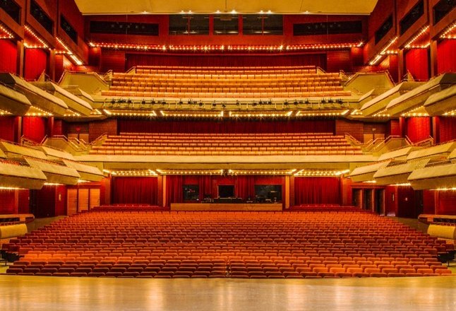 The opulent Thunder Bay Community Auditorium, with shining stage and glowing lighting along the mezzanines, illuminating row after row of red theatre seating. 