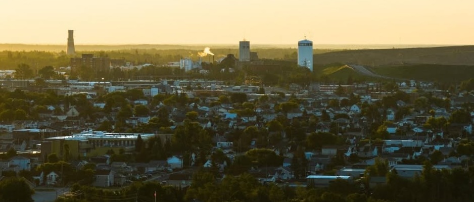 The city of Timmins dotted with lush green trees stretches out under a golden sunset and clear sky.