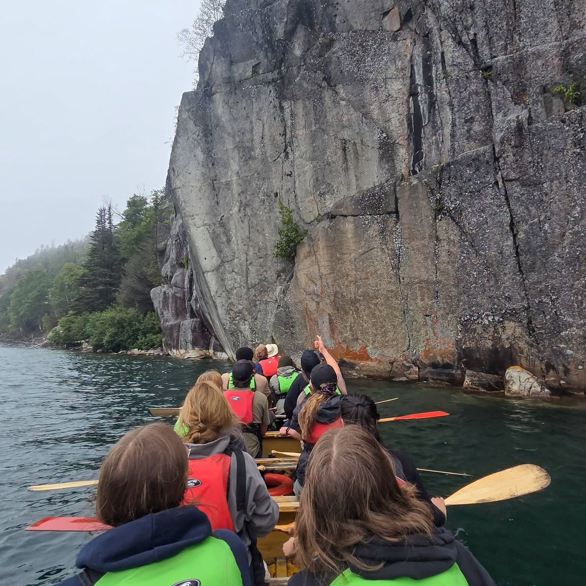 a group of paddlers in a 34 foot canoe stare up at a large flat rock face as they approach on the water of Lake Superior. 