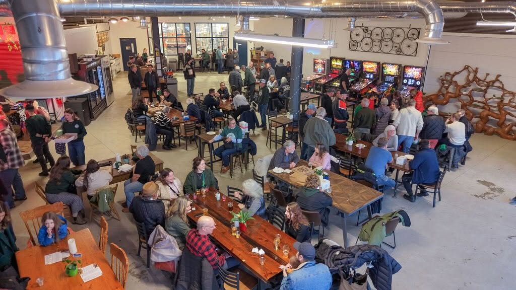 a large crowd gathered around long wooden tables in an open space, brightly lit taproom at the Gateway City Brewery.