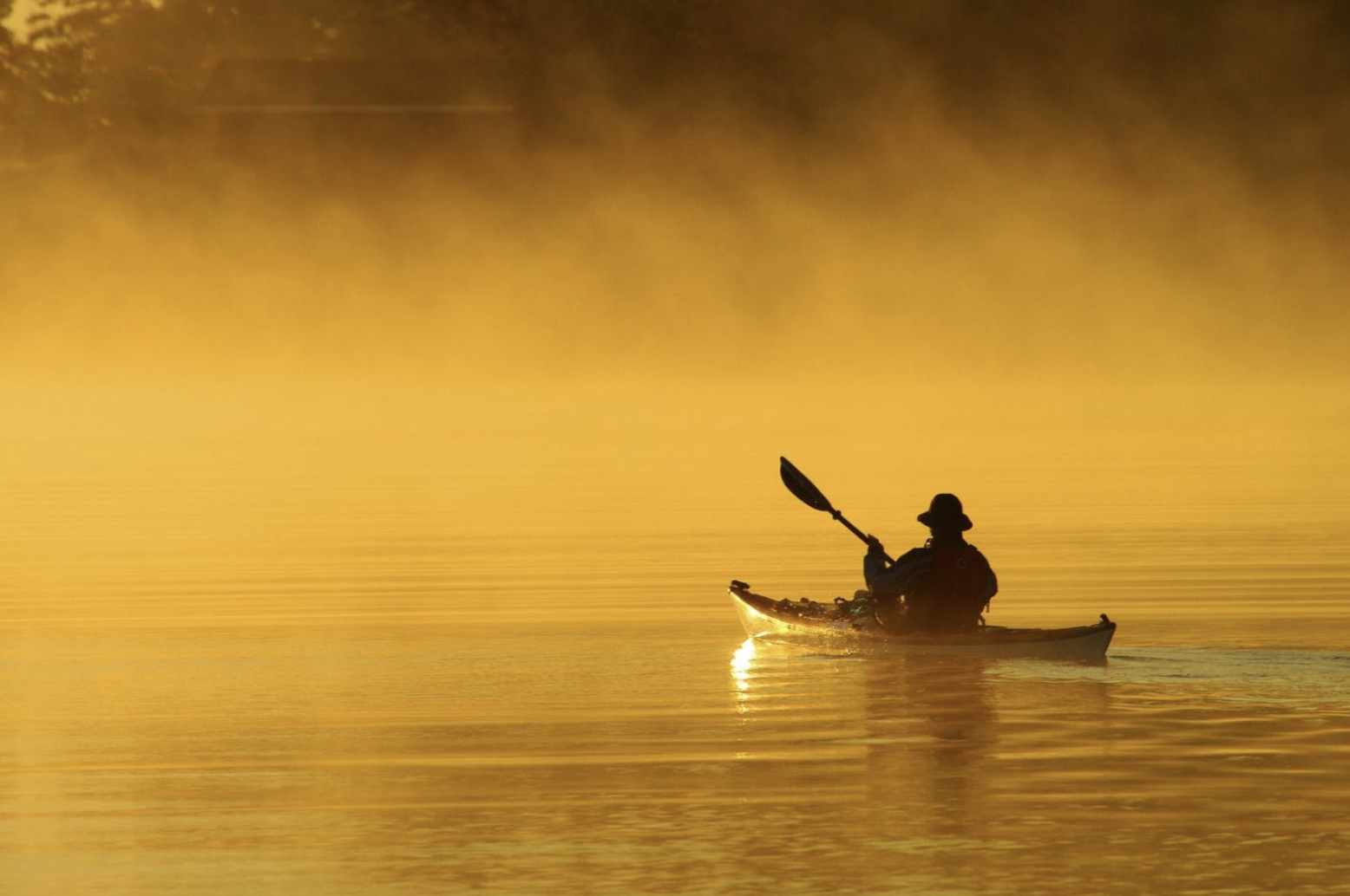 a kayaker paddles out into a misty, glassy lake reflecting a bright golden sunrise. 