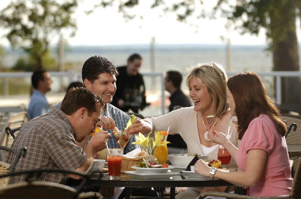 4 people talk and laugh as they share meals at a tree shaded outdoor table near the beach in the summer.