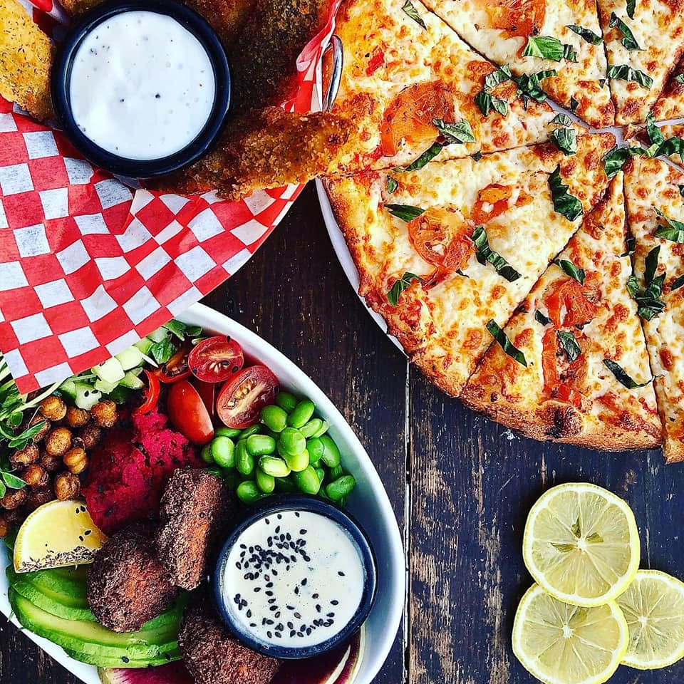 a table set with upscale pizza, chicken fingers and a falafel bowl garnished with an array of fresh vegetables and edamame.