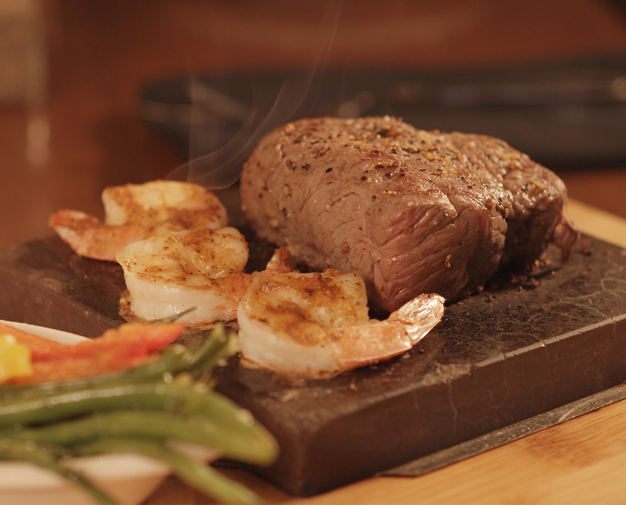 a steaming lava stone with a seared steak and shrimp next to green beans and carrots.