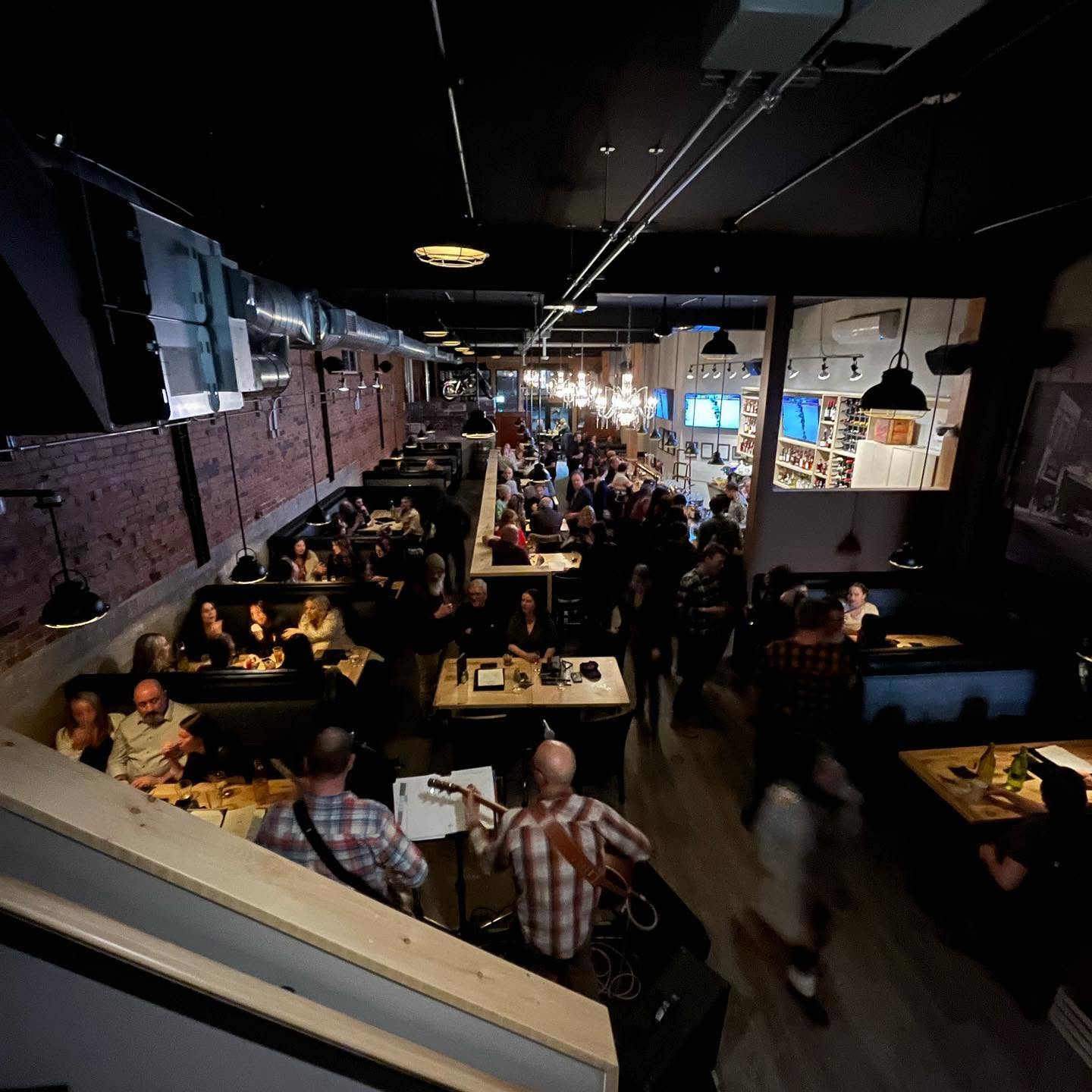 diners sit at rows of tables along a brick and mortar wall in the high-ceilinged Block Public House. A small band plays at the front of the room under soft lighting.