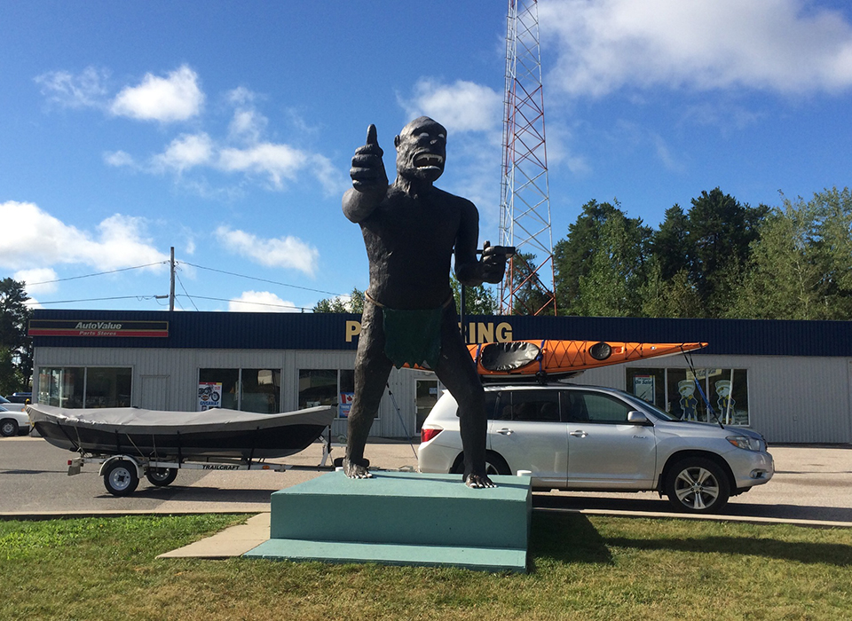 The Vermilion Bay Bigfoot statue, standing on a podium outdoors. It is tall and looming, with a look that's both ferocious and a little comical, flashing a thumbs up sign. 