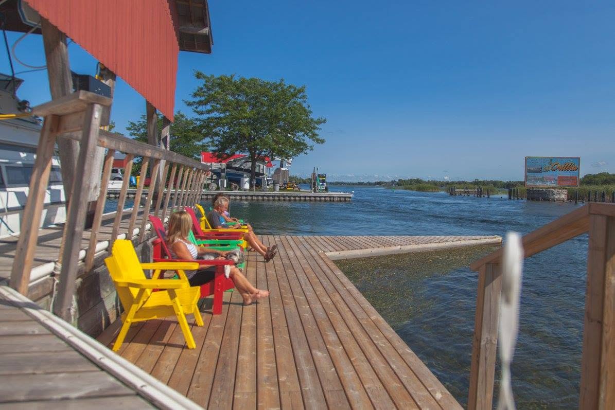 people lounge in colourful wooden deck chairs along the wooden pier in front of the red-roofed fuel station at Bridge Port Marina on a clear summer day. 