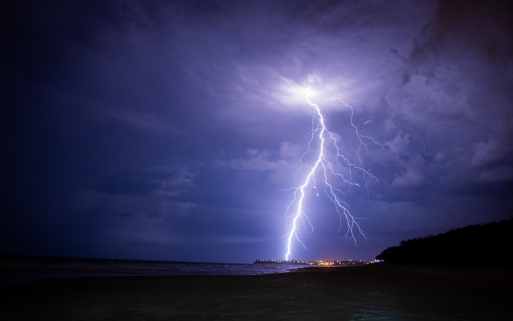 a flash of lightning shoots down to the ground from dark purple clouds in the night sky.