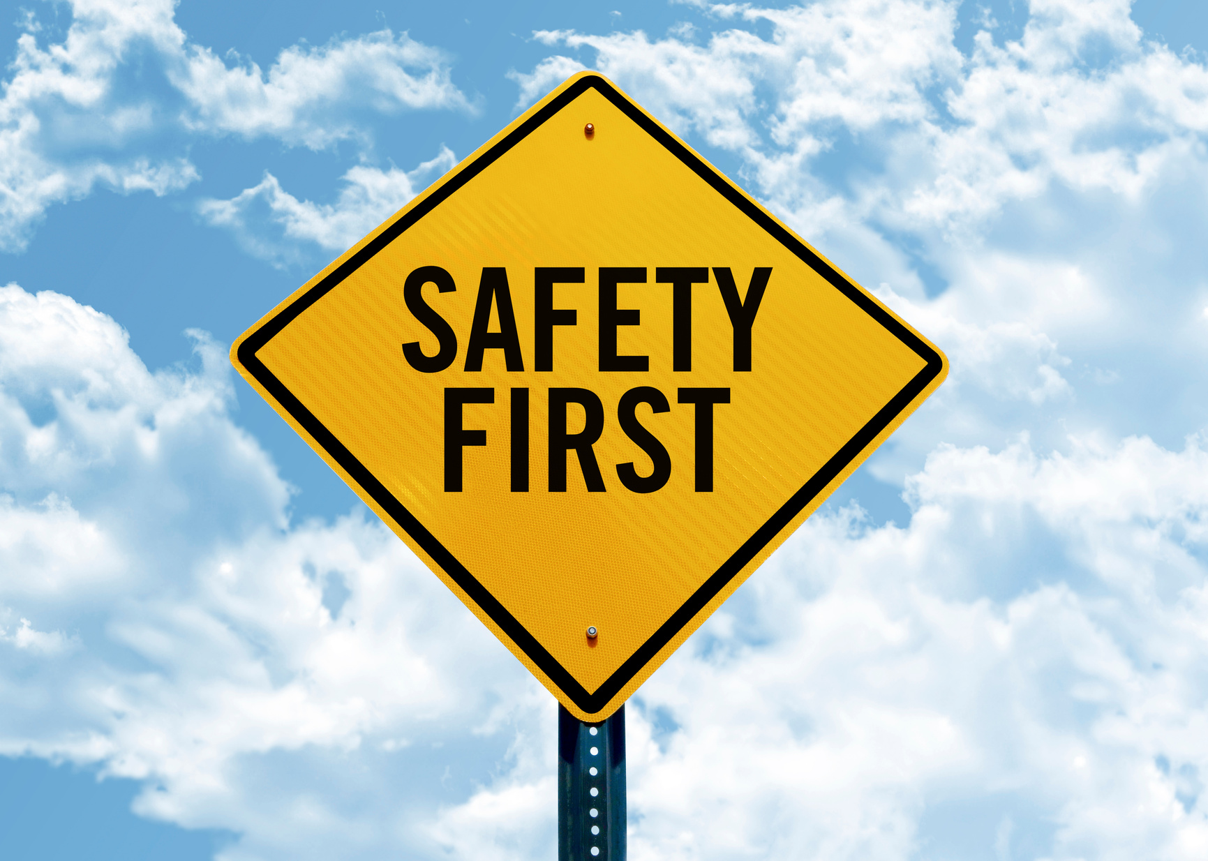 a diamond-shaped yellow caution sign that reads "safety first" against a blue sky with puffy clouds.