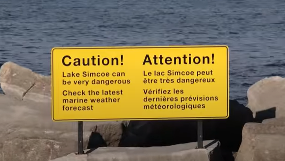 A yellow sign set in the rock on the shore of Lake Simcoe which states "Caution! Lake Simcoe can be very dangerous. Check the latest marine weather forecast."