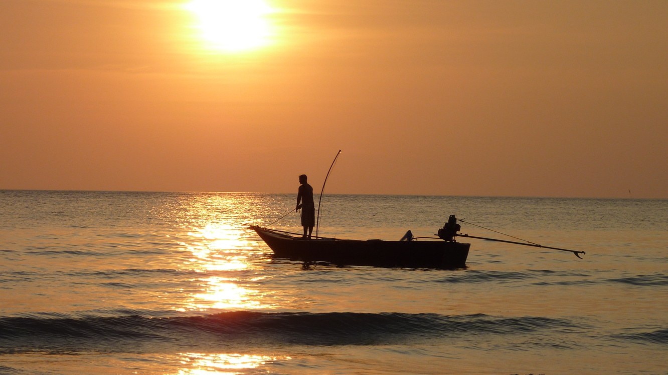 an angler standing in their boat on calm water, silhouetted against a bright golden sunset.
