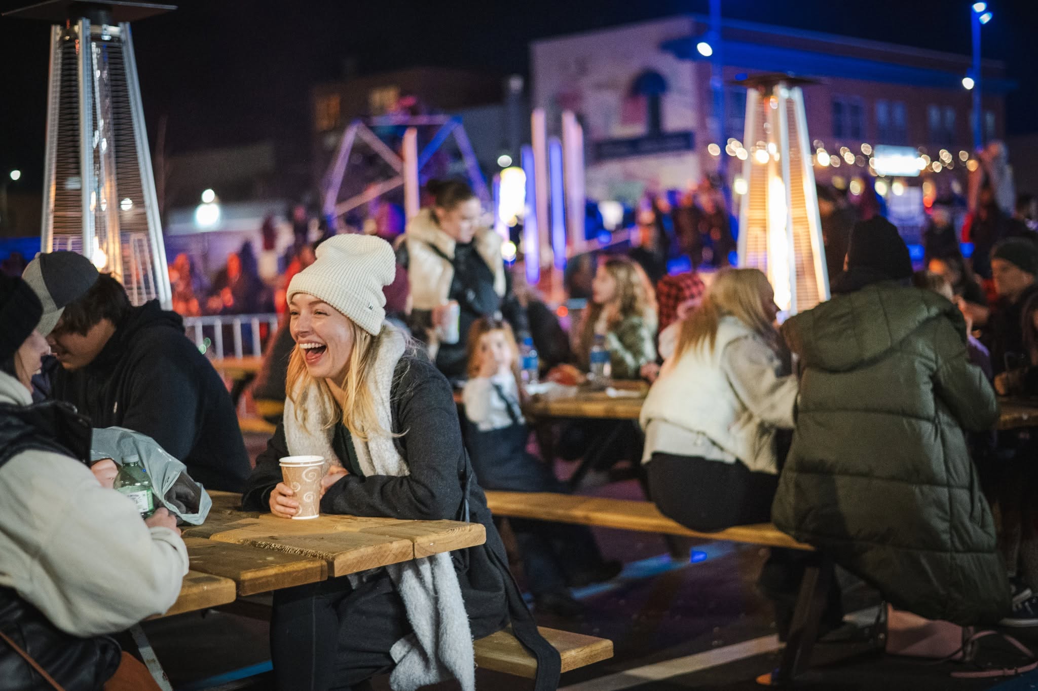 a woman in cozy winter gear with a hot beverage in hand laughs gleefully as she talks with a friend on a busy outdoor patio at night at the Sault Ste. Marie Tree Lighting. 