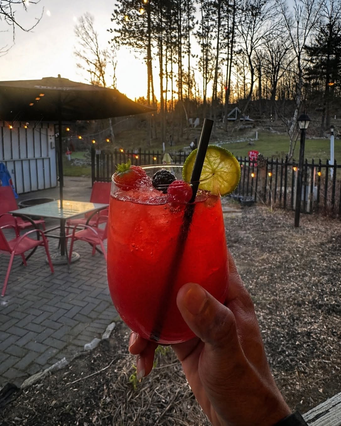 a hand holds up a berry cocktail in front of a forest, trails and a setting sun outside Jolly Roger Inn.