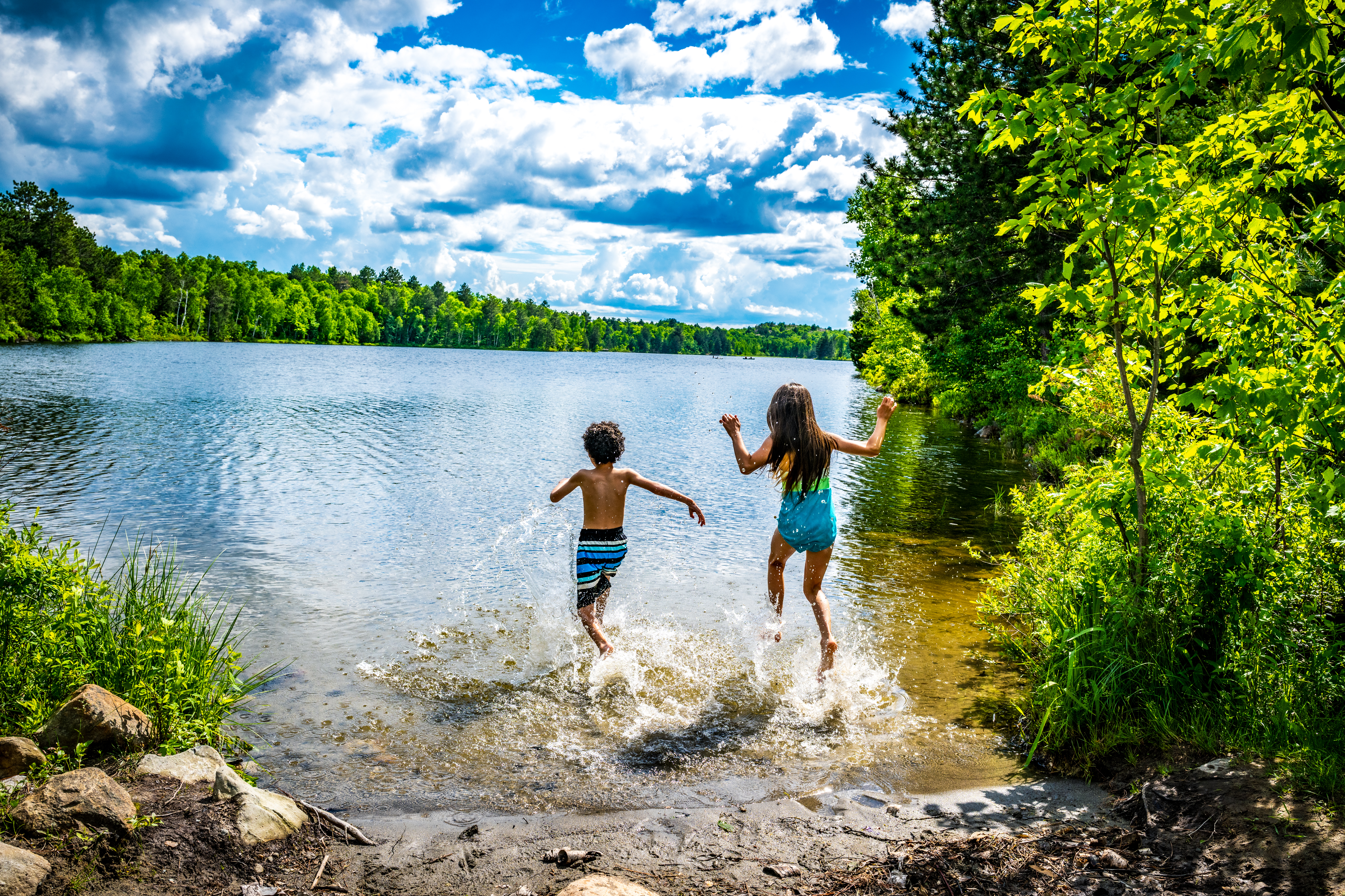 Two children run happily off a beach nestled in the green forest at Crowley Lake, under a bright blue summer sky. 