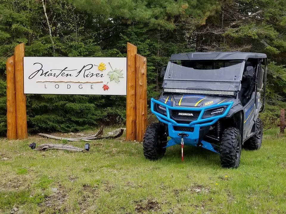 A UTV parked next to a wooden sign that reads "Marten River Lodge" with green trees in the background.