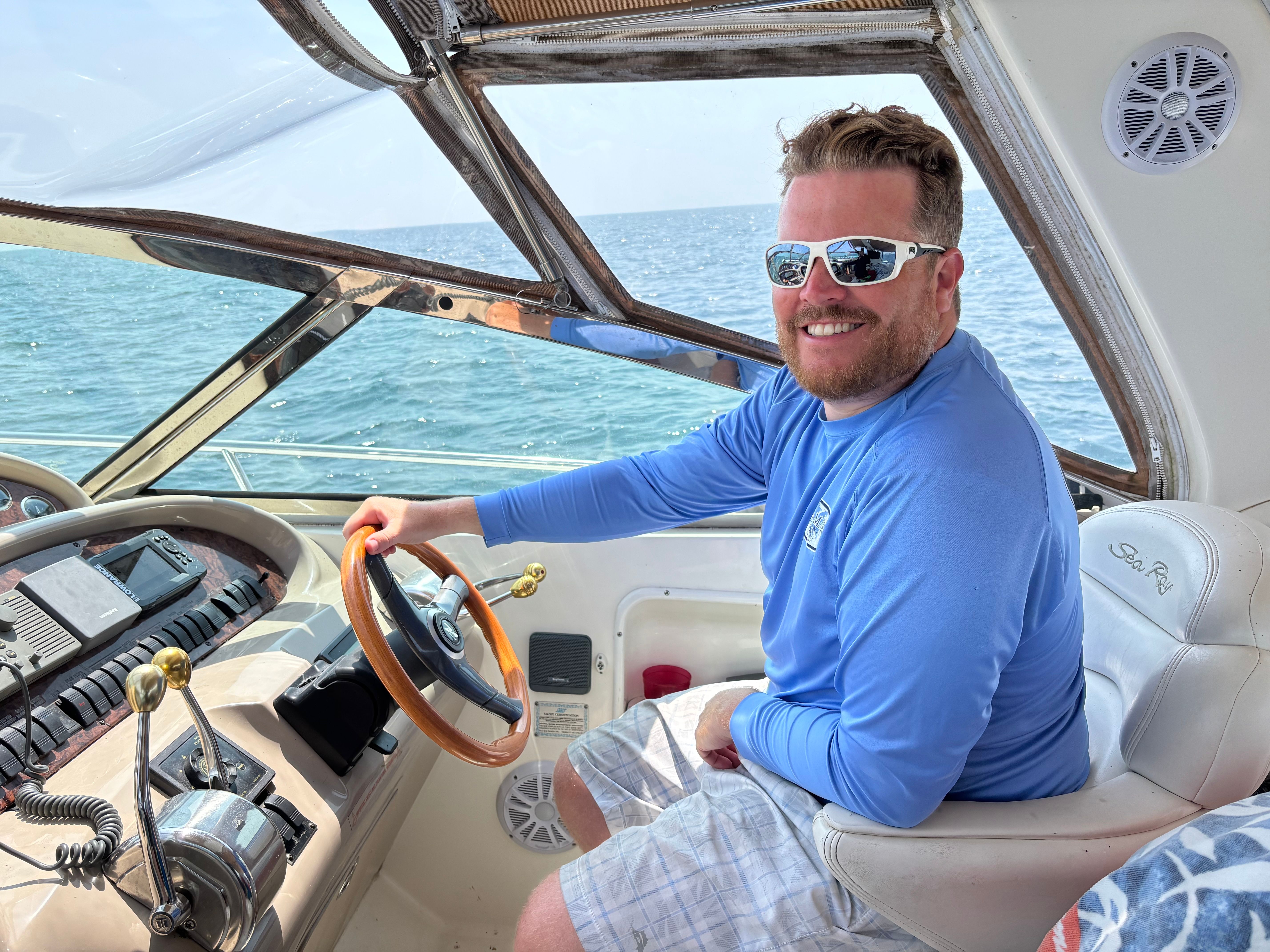 a smiling man wearing sunglasses sitting at the wheel of his boat on a blue lake.