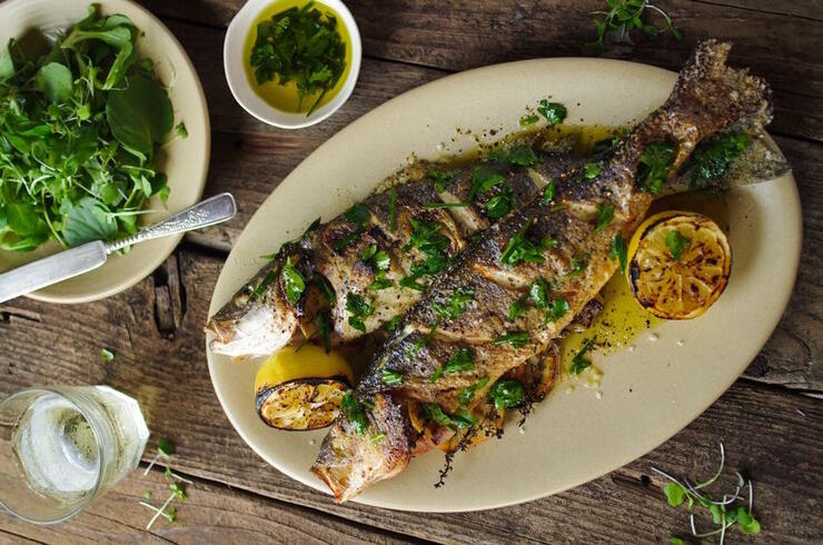 a plate of griled whole trout garnished with herbs and lemon on a wooden table.