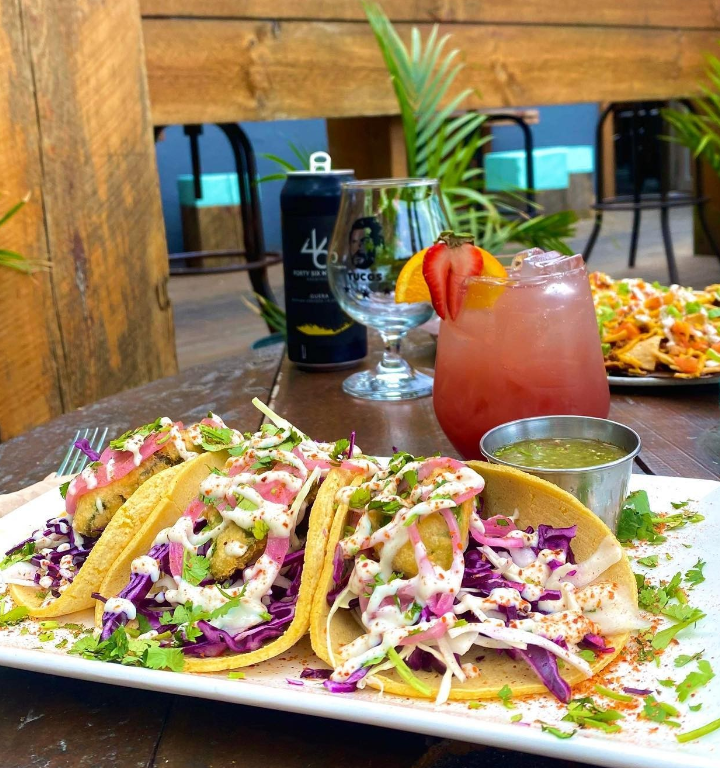 A plate of tacos bursting with fresh vegetables next to a summer cocktail on a wooden table outdoors. 