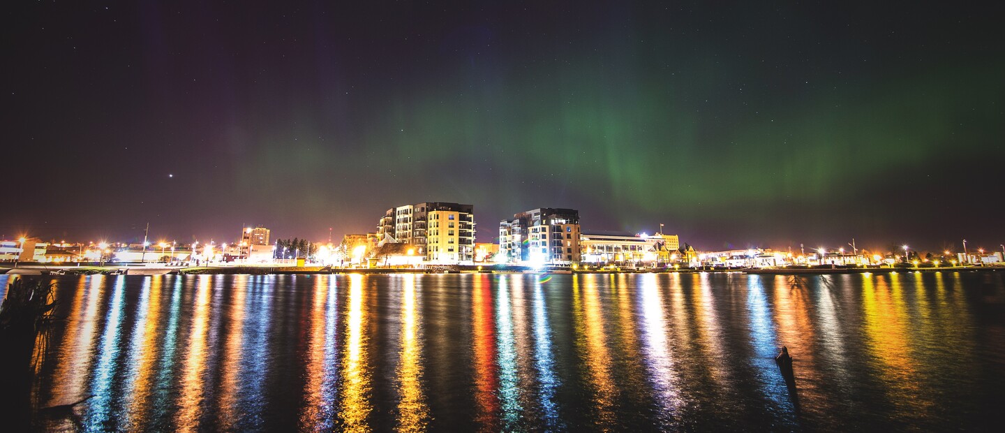 The Thunder Bay skyline from Lake Superior at night, colourfully lit up and reflecting in the glassy water. Green northern lights dance overhead.