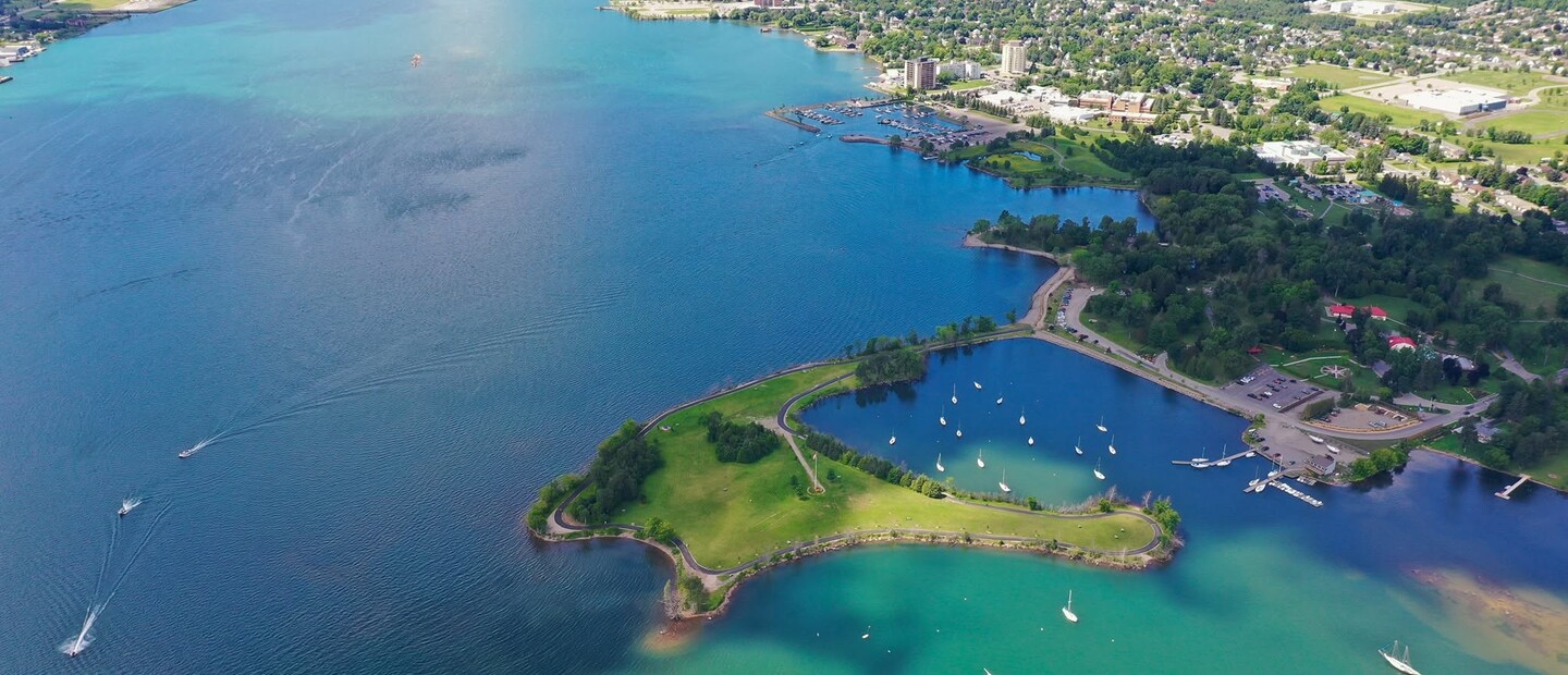 The city of Sault Ste. Marie, Ontario stretches along the shore of the turquoise and blue St. Mary's River, dotted with sail boats next to a lush green shore.