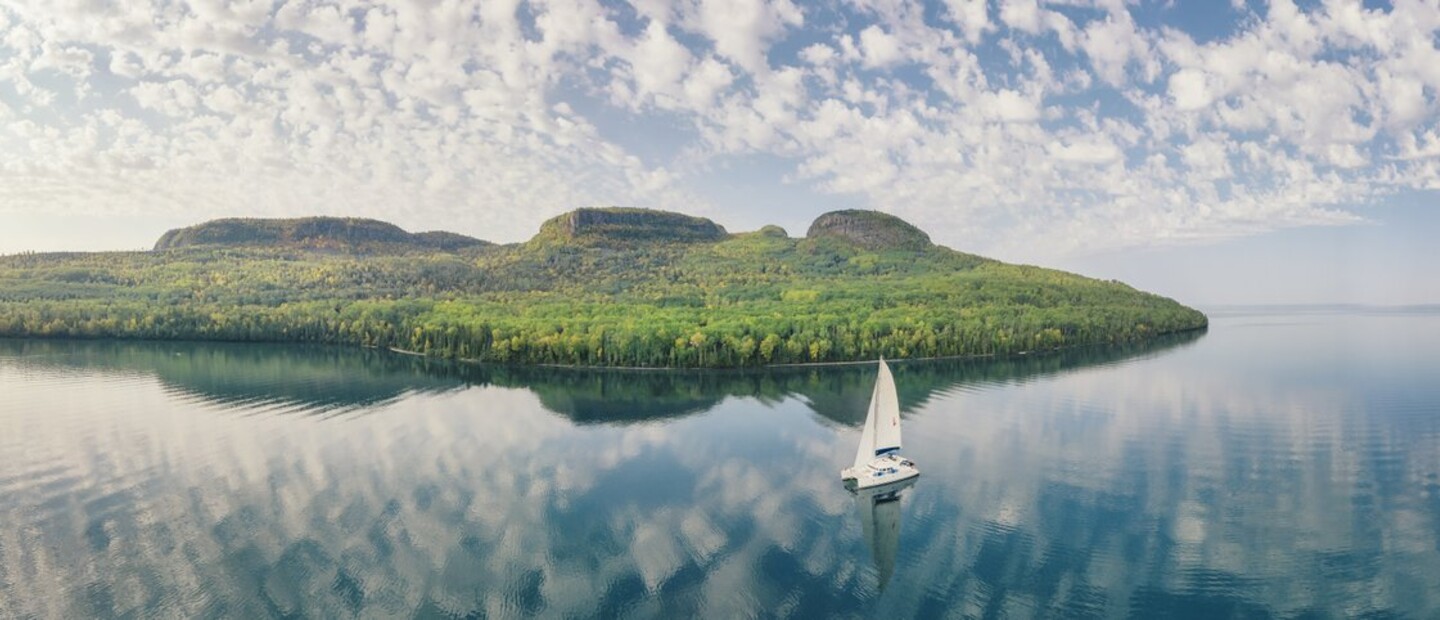 A sailboat floats on glassy Lake Superior, reflecting the summer sunrise and the green banks of Sleeping Giant in the background.