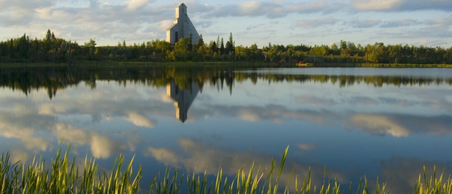 a glassy lakes dotted with green reeds reflects the McIntyre Headframe and a golden sunrise in Timmins. 
