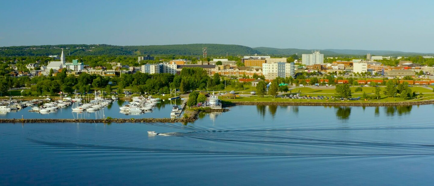 the city of North Bay stretches out along the Lake Nipissing shoreline as a boat glides by on the glassy lake water under a clear blue summer sky. 