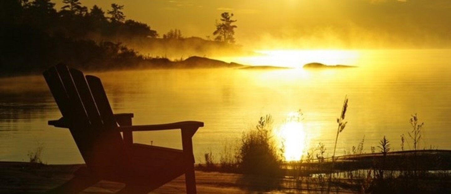 a wooden chair faces out over a lake in the morning, the golden sunrise illuminating the mist on the water.